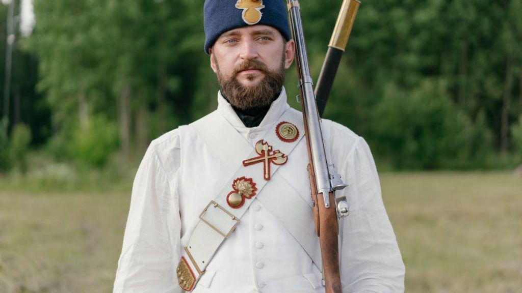 Man in historical military uniform with rifle, outdoors in summer.
