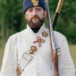 Man in historical military uniform with rifle, outdoors in summer.